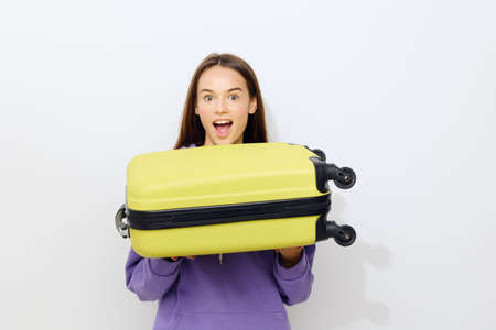 an admiring beautiful woman stands on a white background in a purple tracksuit holding a suitcase from below with two hands smiling broadlyの写真素材