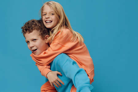 cute, beautiful, happy, playful children, a boy and a girl of school age stand on a blue background in bright plain clothes, sideways to the camera and the boy rolls the girl on his backの写真素材