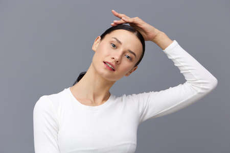 a cute woman with a ponytail on her head stands on a dark background in a white tight T-shirt, put her hand on her head, turning asideの写真素材