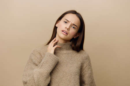 a frustrated, sad, beautiful woman in a warm, textured sweater stands on a beige background touching her neck with her fingers and looks at the camera. Horizontal Studio Photographyの写真素材