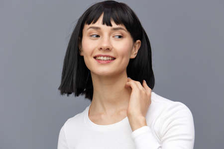 a relaxed woman stands against a dark background in a tight white T-shirt, calmly looks forward with her hand on her neckの写真素材