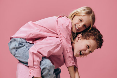 funny, happy kids brother and sister play and the boy rolls the girl on her back standing sideways to the camera in pink clothes on a pink background. Relationships, friendship, familyの写真素材