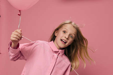 happy, joyful girl of school age, blonde, stands playing with a pink balloon in her hand and smiles joyfullyの写真素材