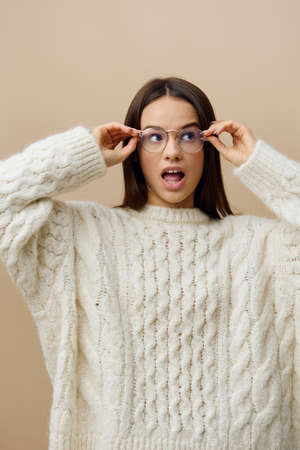 a happy, mischievous woman in eyeglasses stands with her mouth wide open with happiness and holding her glasses with her hands while standing on a beige background.の写真素材