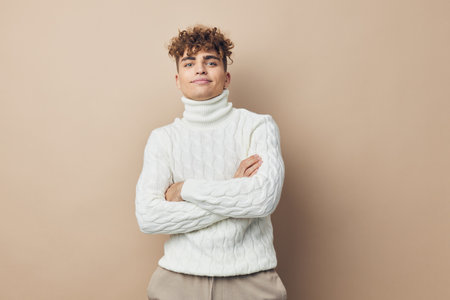 handsome, pleasant man with curly hair, standing in a white knitted sweater on a beige background and smiling pleasantly with his arms crossed on his chest. horizontal studio photo with an empty spaceの写真素材