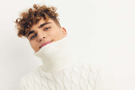 a close photo of a handsome, attractive man with curly hair, standing in a white knitted sweater on a bright background, smiling pleasantly into the camera with a tight neck on his faceの写真素材