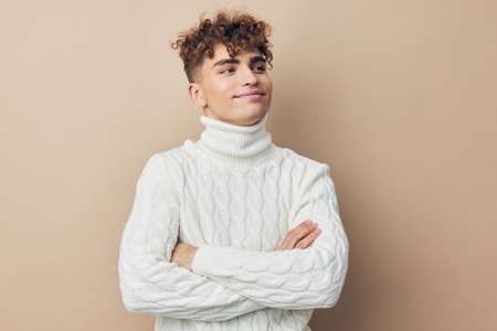 handsome, pleasant man with curly hair, standing in a white knitted sweater on a beige background...