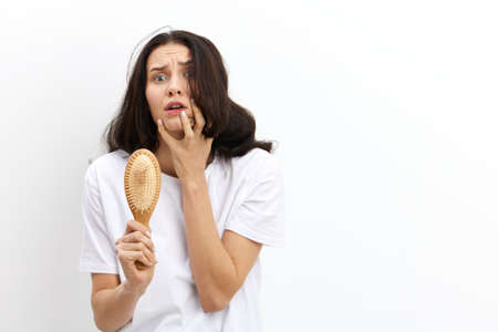 horizontal photo on a white background of a sad, shocked woman holding a wooden comb in her hands and clutching her hair in panic, looking into the camera with her eyes wide openの写真素材