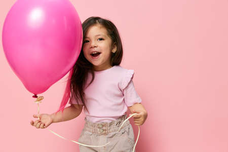 cute, happy little girl stands with a big pink balloon on a pink background in fashionable, stylish clothes and smiles broadly with happiness looking at the cameraの写真素材