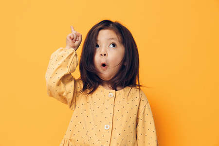an adorable, playful little girl in a summer dress stands on an orange background with an emotional, very surprised face and points her index finger upの写真素材