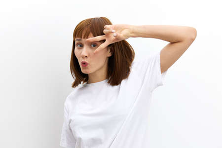 a mischievous, cheerful, pleasant woman with red hair standing on a white background with an empty space for inserting an advertising layout and showing a victory sign near her face with a gestureの写真素材