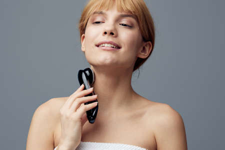 a woman with perfect skin stands on a gray background wrapped in a towel and massages her neck with a black electric anti-aging massager, smiling pleasantly with her eyes closed.の写真素材
