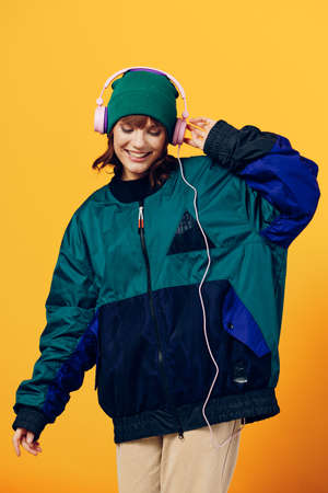 a happy, joyful woman in a green jacket and hat enjoys music standing in pink headphones on a yellow...