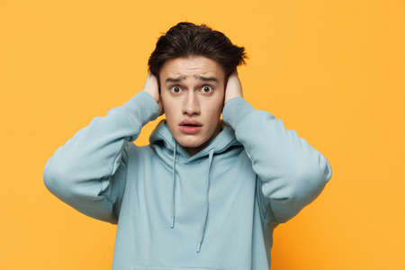 a close horizontal photo of a shocked young man standing on an orange background in a light blue hoodie, covering his ears with his hands, and looking very emotionally into the cameraの写真素材