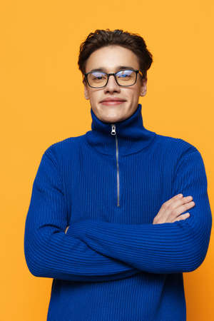an attractive young man stands on an orange background in a blue zip-up jacket and black eyeglasses...