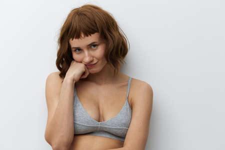 a funny, slightly upset tanned woman with red hair stands on a light background in a gray top and supports her cheek with a fist of one handの写真素材