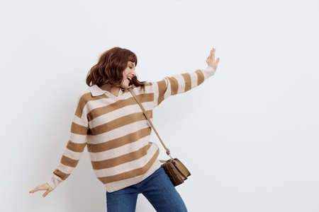 a happy woman standing on a white background in a striped sweater stood half sideways with her hands in different directions and hanging her bag on one shoulder, looking awayの写真素材