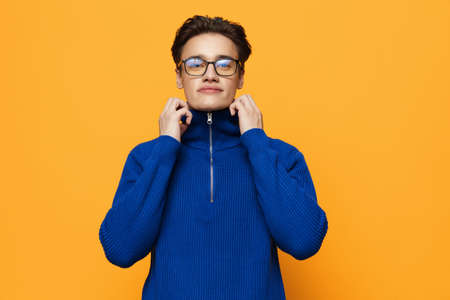an attractive young man stands on an orange background in a blue zip-up jacket and black eyeglasses and pulls aside the neck of the jacketの写真素材