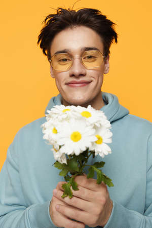 a close vertical portrait of a funny man standing on an orange background in a light blue hoodie, wearing yellow glasses, holding a bouquet of white daisies and smiling pleasantlyの写真素材