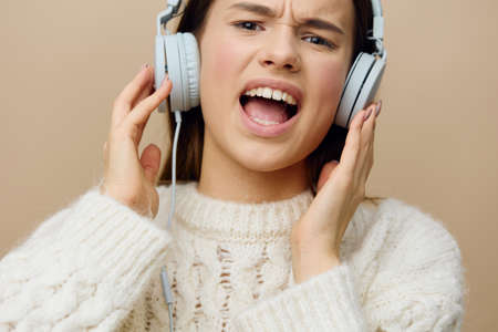 close horizontal photo of a woman in a white sweater listening to music in her wired headphones smiling broadly with happiness and holding them with her hands while looking at the camera with delightの写真素材
