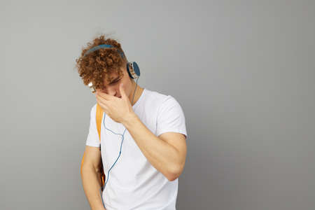 a student guy stands on a gray background in a white T-shirt and listening to music with headphones...