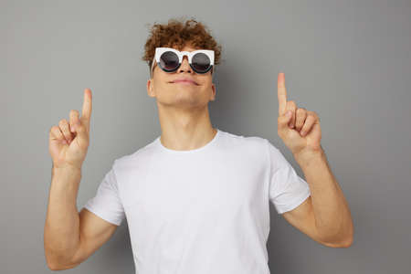 a young man stands in a casual T-shirt with funny glasses on his face and holds his hands up, pointing his fingers up. Horizontal photoの写真素材