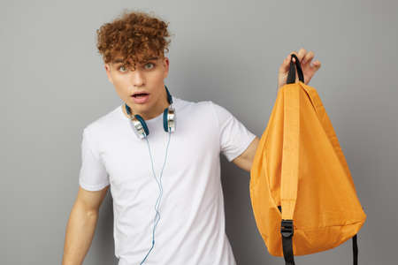 the student stands on a light gray background with headphones around his neck and looks at the camera with delight, holding his orange backpack in his handの写真素材