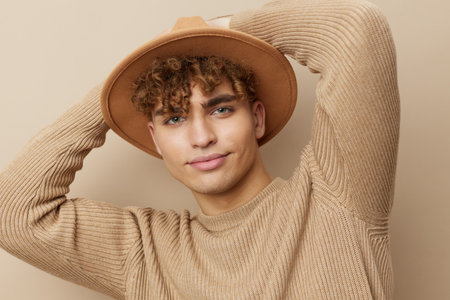 a handsome young man stands on a beige background in a stylish sweater with a hat on his head and...