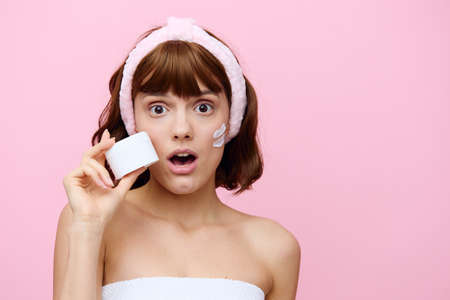 a cute young woman stands on a pink background in a white towel with cream applied to her cheek,...
