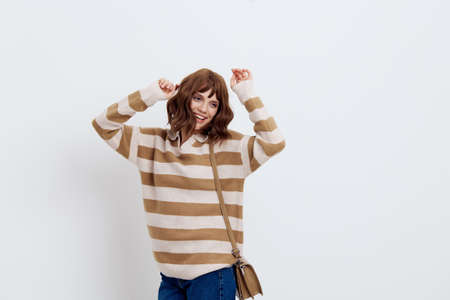 a beautiful, cheerful woman stands on a white background in a stylish striped sweater with a beige bag on her shoulder and smiles cheerfully while standing in a relaxed pose, raising her hands upの写真素材