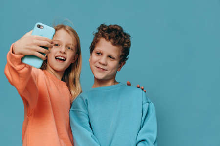 cute, beautiful children, brother and sister stand in bright clothes on a blue background and the girl takes a selfie on her smartphone hugging her brotherの写真素材