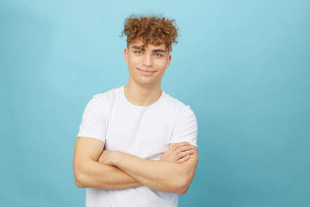 an attractive man with curly hair stands on a light blue background in a white cotton T-shirt and looks at the camera with his arms crossed on his chest. Horizontal studio photography with empty spaceの写真素材
