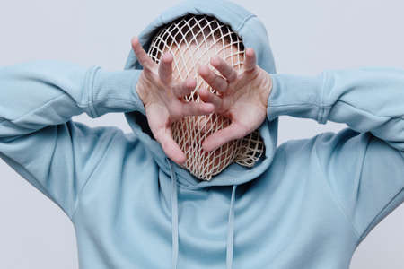 horizontal studio photo of a strange, tortured man in a light blue hoodie covering his face with his...