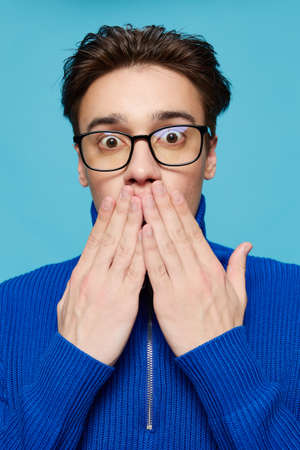 portrait of a handsome man in a blue zip-up sweater and black eyeglasses, standing on a light blue background looking into the camera covering his mouth with his handの写真素材