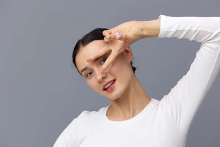 a sweet, beautiful, attractive woman smiles sweetly looking at the camera standing on a gray background in a white T-shirt showing a victory sign near her face with her fingers.の写真素材