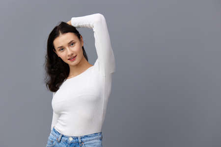 an excited woman stands on a gray background in a white T-shirt, raised her elbow above her head,...