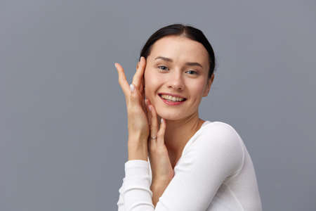 a cute woman with a ponytail on her head stands on a dark background in a white tight T-shirt, put two hands on her cheek and smiles happilyの写真素材