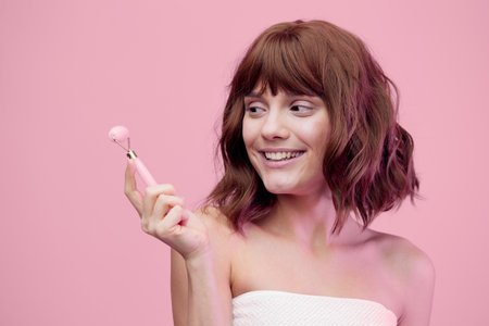 a happy, enthusiastic woman, standing on a pink background with pink lighting from the side, with her hair styled, holding a roller massager in her hand, squinting her eyes with pleasure.の写真素材