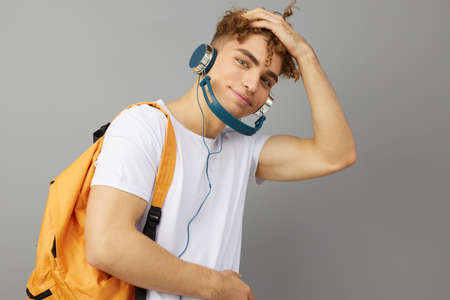 a puzzled student guy stands on a gray background in a white T-shirt and listening to music with...