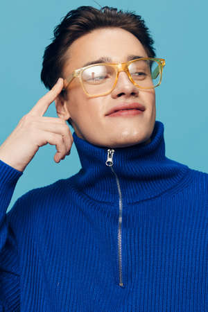 a cute, handsome man stands on a light blue background in a blue zip-up sweater and happily smiling...