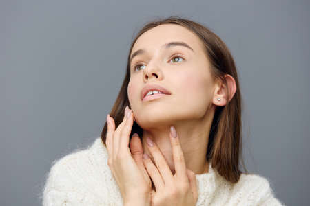 close horizontal portrait of a graceful tender woman looking up with her hands near her face in a white sweaterの写真素材