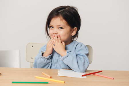 a cute, pleasant little girl of pre-school age sits at a table and draws with colored pencils, smiling covering her face with her hands. Themes of hobby, development and trainingの写真素材
