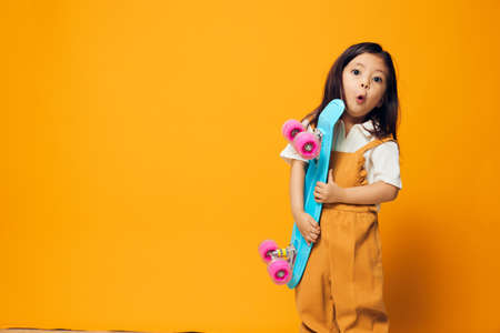 a cute, adorable little preschool girl stands with a blue skateboard in her hand, smiling adorably at the camera. Horizontal photo with empty space to insert an advertising layoutの写真素材