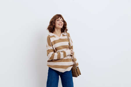 cheerful woman stands on a white background in a stylish striped sweater with a beige bag on her...