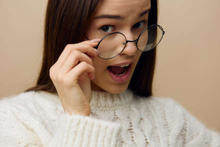 a pretty woman stands on a light background in a white sweater and holds her black glasses with her hands, turning her head and looking suspiciously at the front, wrinkling her foreheadの写真素材