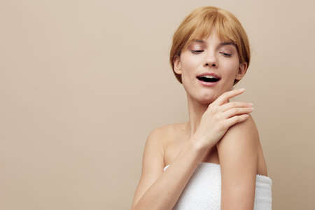 a sweet, elegant woman stands on a beige background wrapped in a white towel and smiling pleasantly with her eyes closed gently touches her shoulderの写真素材