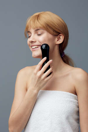 a happy, joyful woman stands on a gray background wrapped in a towel and does a facial massage with a black electric massager, covering her eyes with pleasureの写真素材