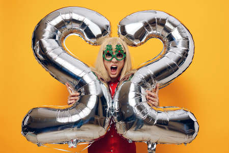 a happy emotional woman stands in a red shirt with green festive glasses, holding balloons in the...