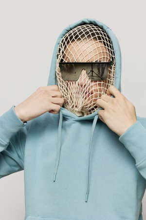 a close studio photograph of a man in a light blue hoodie, a mesh stretched over his face, standing on a light background, with sunglasses covering his eyes, holding the edge of the hoodieの写真素材