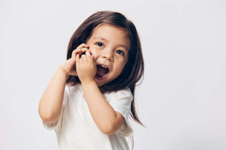 a little girl of preschool age, sitting on a white background in a white T-shirt, enthusiastically talking on the phone, turning half-side to the camera. The topic of the conversation on the phoneの写真素材